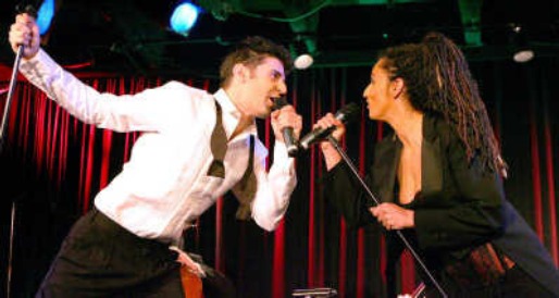 Roger Stein und Sandra Kreisler singen einander mit Mikrofonen zu, vor rotem Vorhang