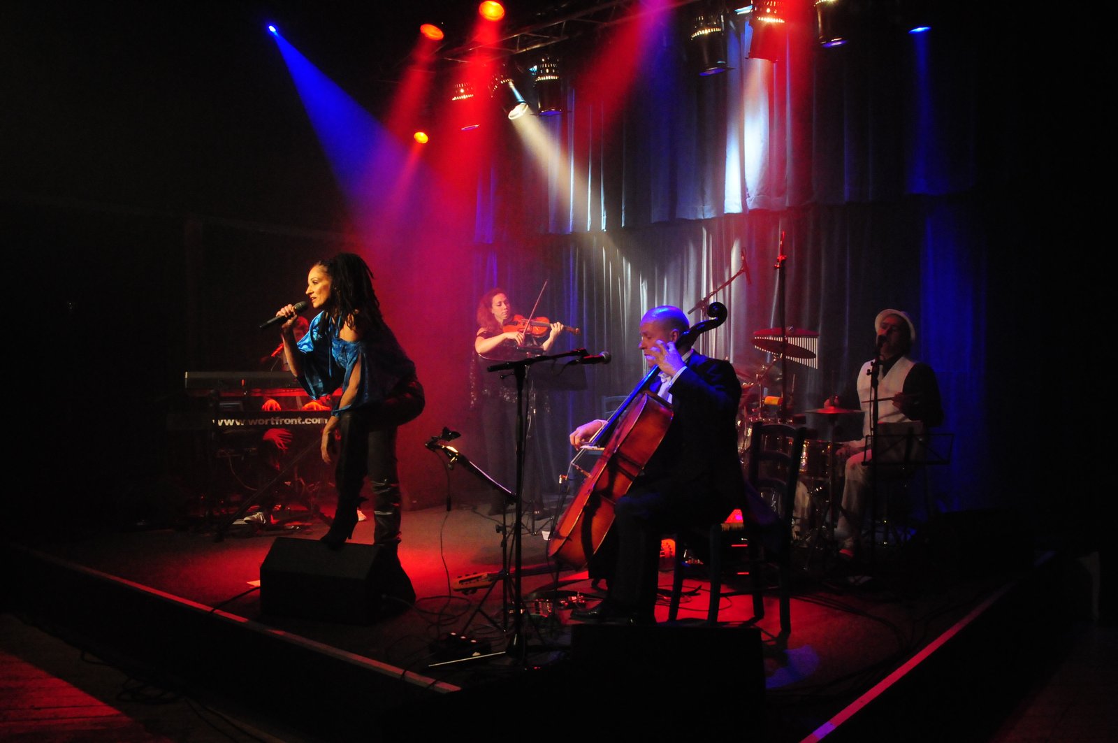 Wortfront auf der Bühne in buntem Licht: Sandra Kreisler am Mikrofon, Roger Stein am Keyboard, Anna Eichholz mit Geige, Ulrich Maiss am Cello und Paolo Eleodori am Schlagzeug
