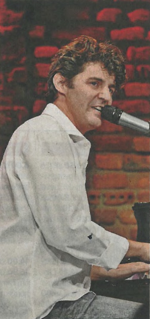 Roger Stein im weißen Hemd am Flügel, vor einer roten Backsteinwand