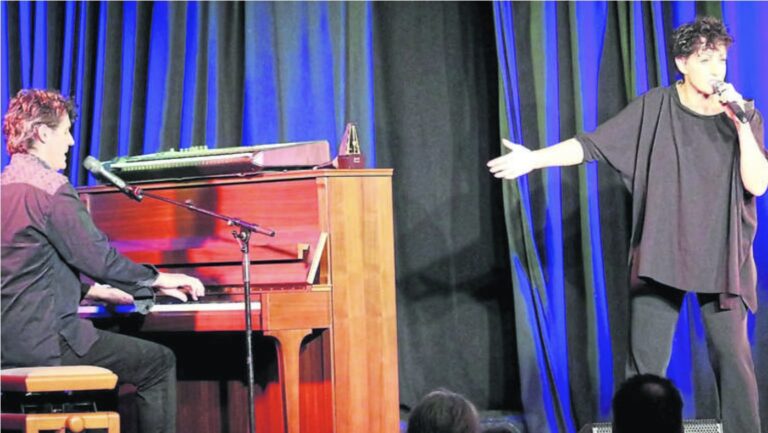 Roger Stein am Klavier links, Sandra Kreisler rechts mit Mikrofon und ausgestrecktem Arm vor blauem Vorhang
