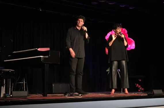 Roger Stein und Sandra Kreisler stehen mit Mikrofonen auf der Bühne, links ein Flügel, rechts eine große pinke Blume